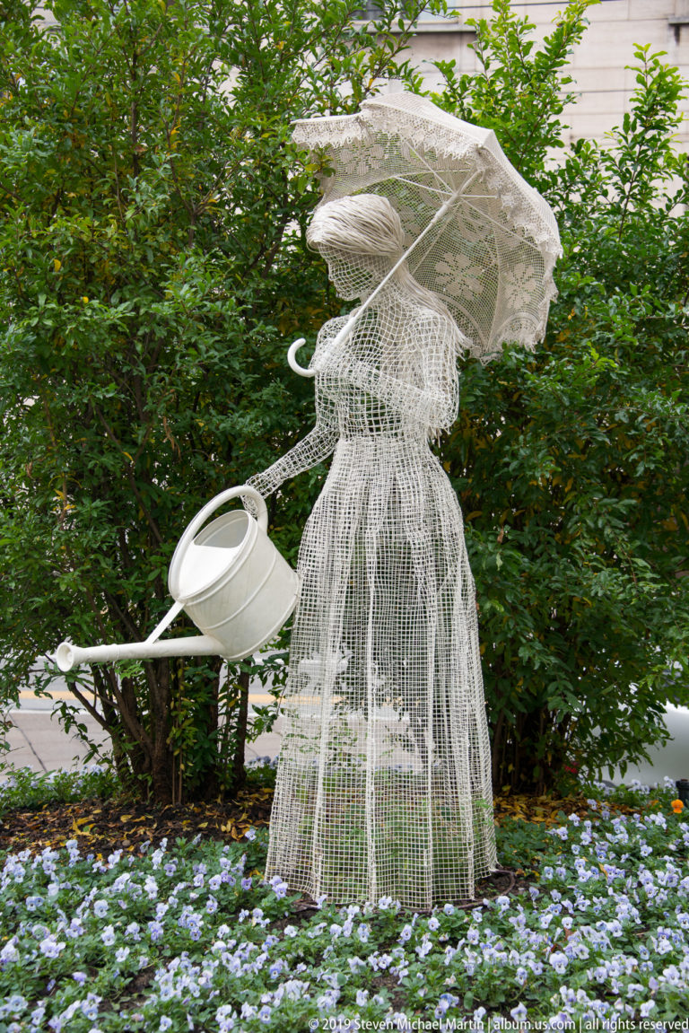 White wire-frame woman with a broad hat and watering can standing among flowers and shrubs in a Turin garden.