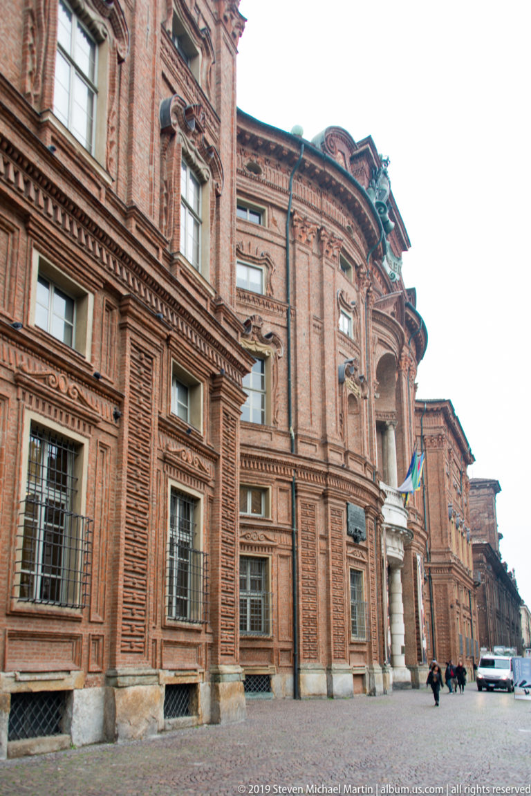 Oblique street view along Palazzo Carignano’s textured red-brick façade toward its ornate entrance.