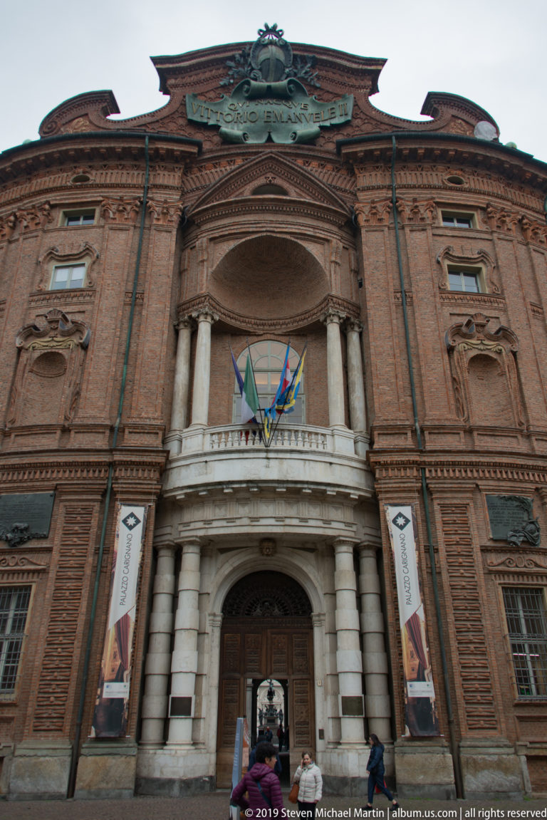 Central entrance of Palazzo Carignano beneath flags, framed by richly modeled red-brick Baroque architecture.