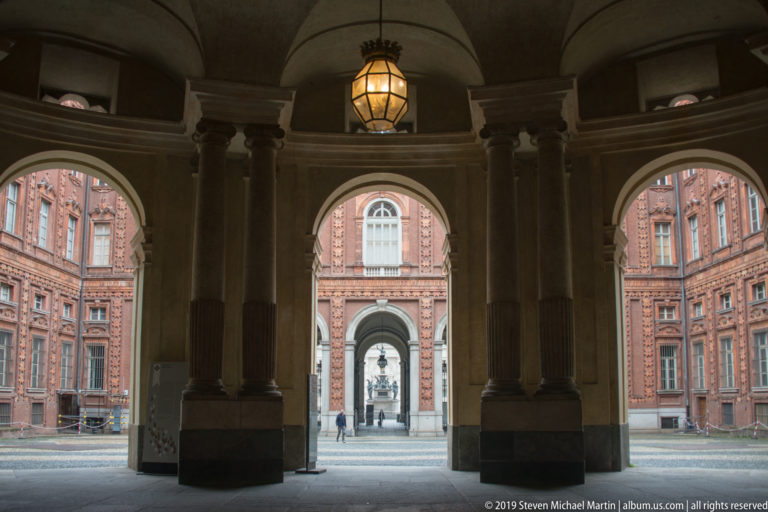 Shadowed vaulted portico with columns, hanging lantern, and aligned arches opening into Palazzo Carignano’s courtyard.