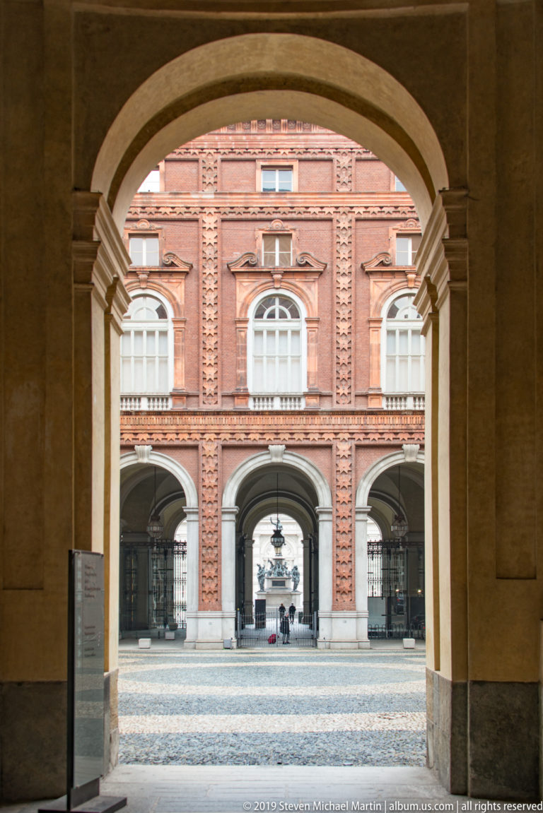 Stone archway framing Palazzo Carignano’s brick courtyard façade and a second distant arch.