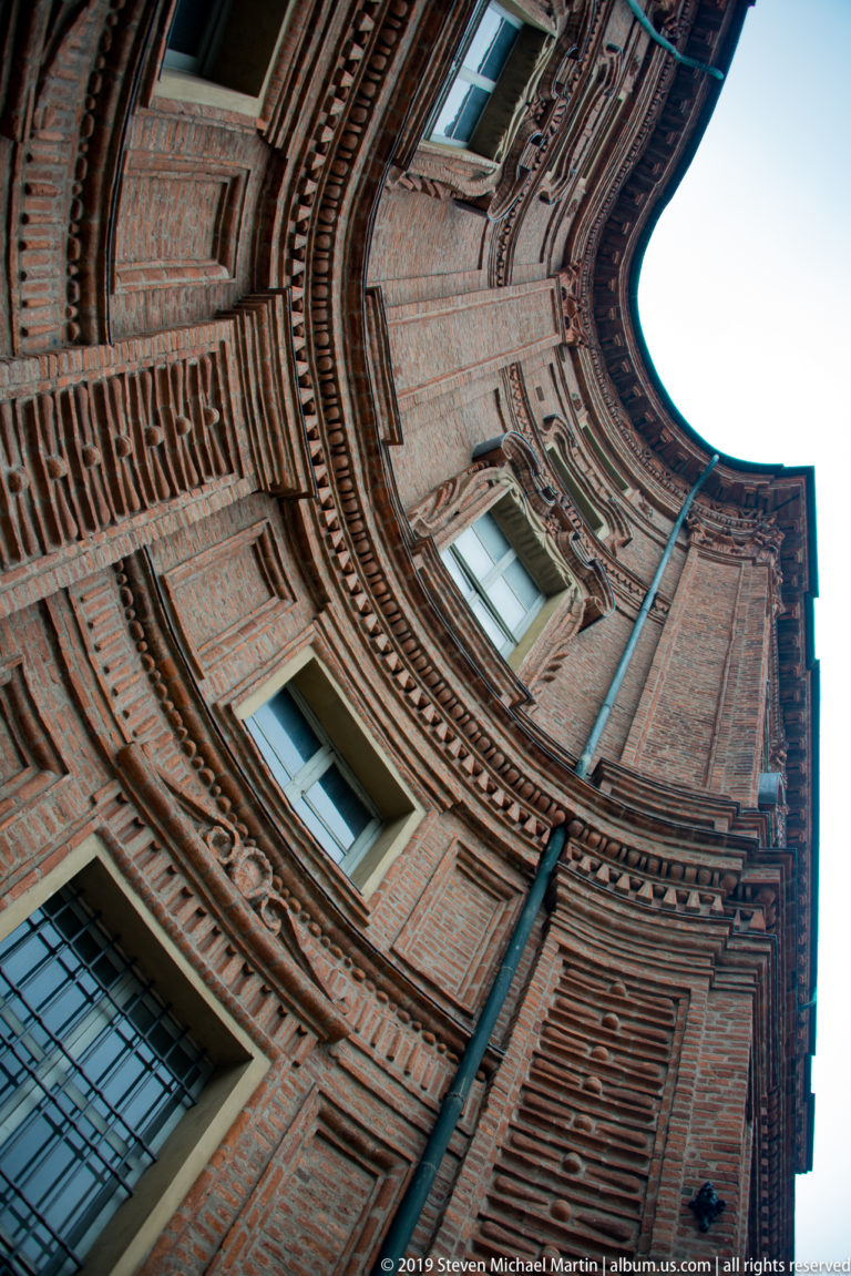 Steep upward view between curved red-brick walls and windows of Palazzo Carignano, with a narrow opening of sky.