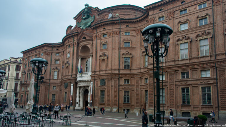 Curving red-brick façade of Palazzo Carignano with ornate stone entrance, windows, and street lamps.