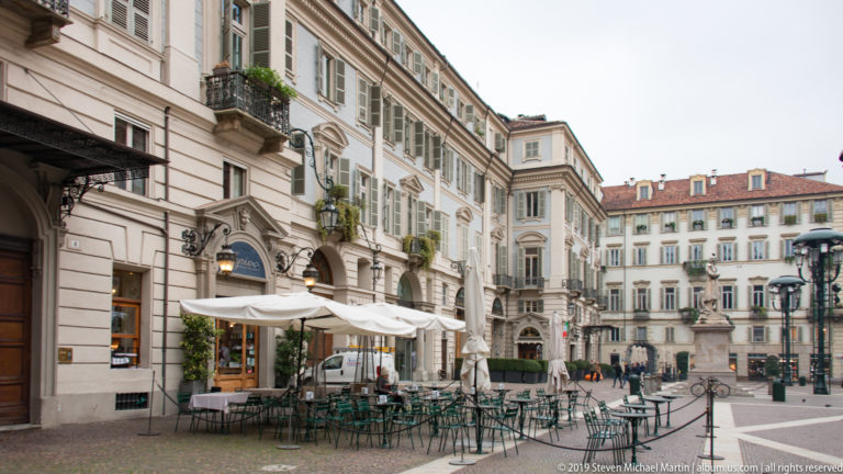 Outdoor café tables beside pale historic façades on a pedestrian street in central Turin.