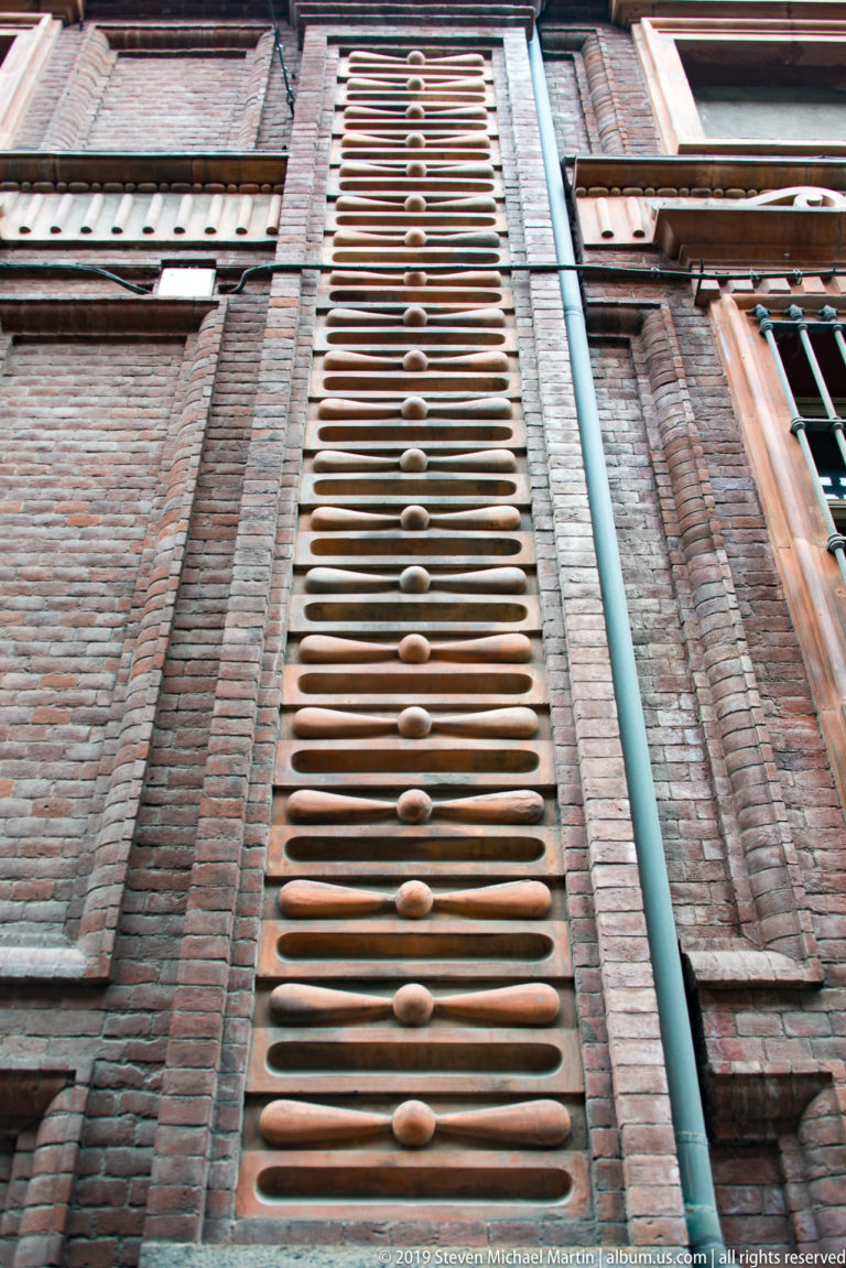 Close upward view of patterned brick courses with rounded projections, a drainpipe, and window edges.