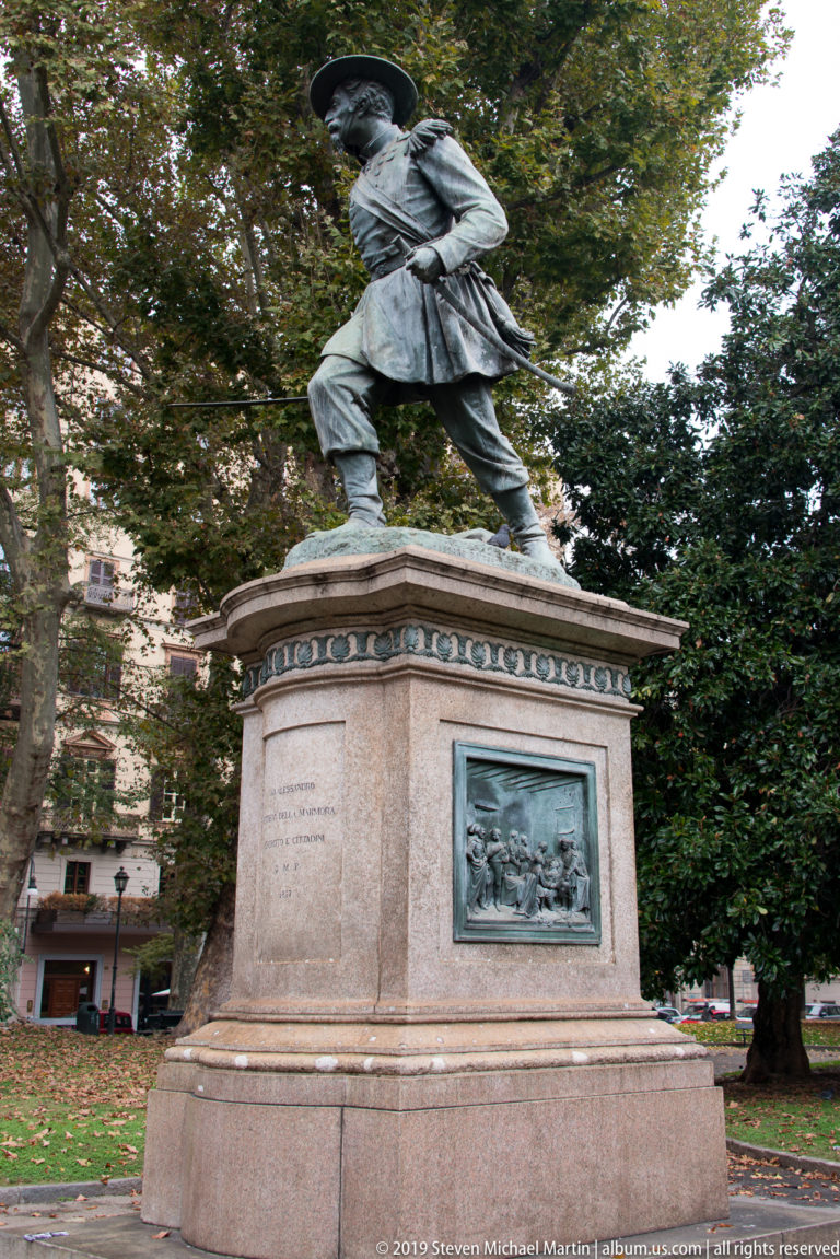 Standing male statue on a tall stone pedestal with a bronze relief panel, surrounded by trees in Turin.