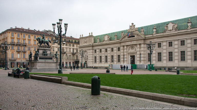 Wide view across Piazza Castello with an equestrian monument, lawn, pedestrians, and a long historic palace façade.