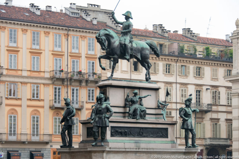 Bronze equestrian monument with standing and seated figures around its pedestal in a Turin square.