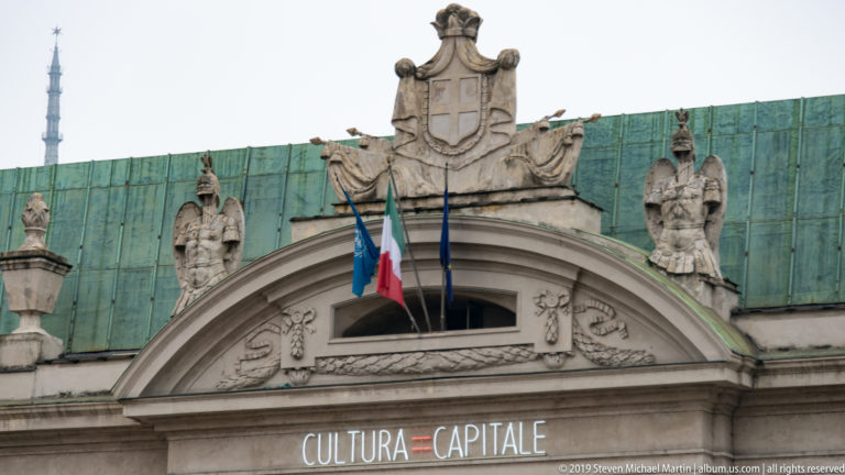 Carved pediment and rooftop figures above flags and the illuminated words “CULTURA = CAPITALE.”