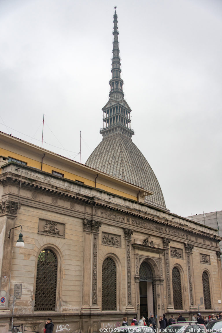Mole Antonelliana dome and tower rising behind a long historic masonry building with arched windows.
