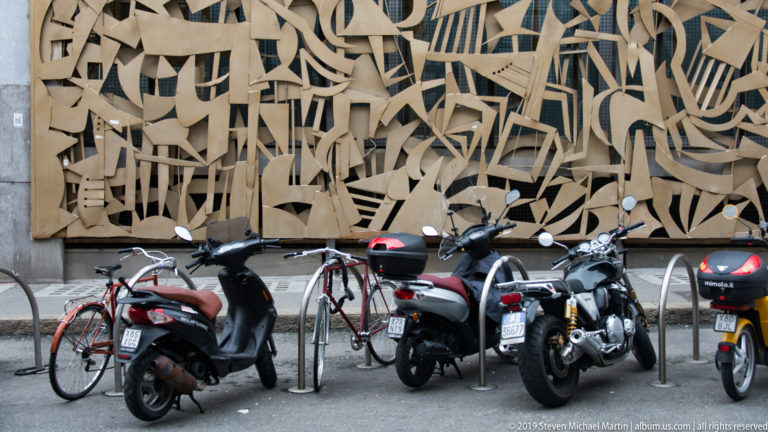 Parked bicycles and motor scooters in front of a large abstract wall relief in Turin.