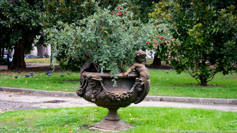 Ornate dark metal garden urn with small child figures, garlands, lawn, and leafy trees in a Turin park.