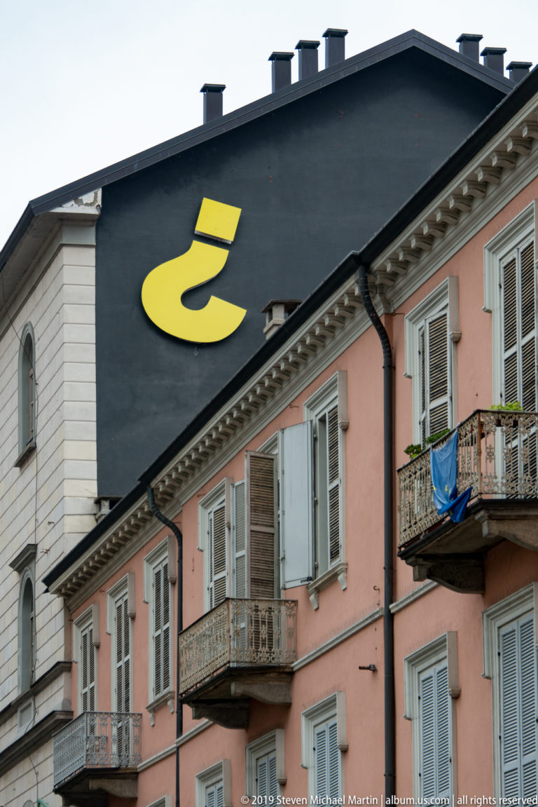 Large yellow question-mark-like symbol painted on a dark gable above pastel residential façades and balconies.