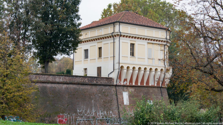 Pale pavilion with tiled roof above a high brick wall, surrounded by autumn trees and vegetation.
