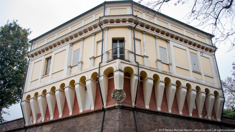 Pale corner pavilion above a brick wall, with repeated arches, red triangular panels, and a circular stone medallion.