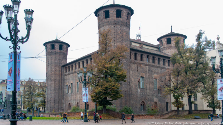 Brick Castello del Valentino with round corner towers, conical roofs, trees, and pedestrians along the street.
