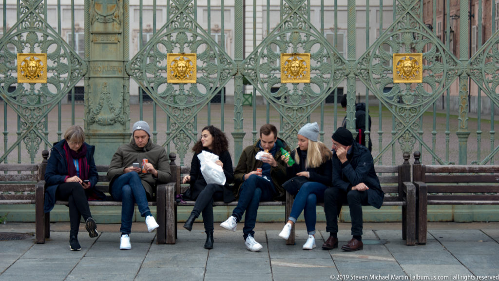 Six people seated on benches, eating and checking phones in front of an ornate green iron gate with gilded emblems.