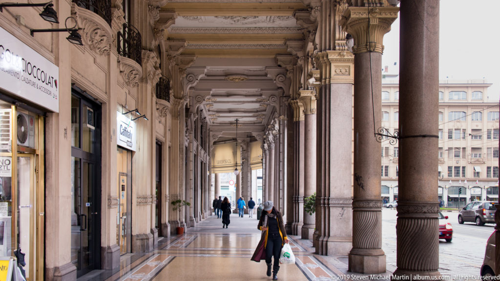 Long Turin portico with stone columns, ornate capitals, shops, pedestrians, and a street visible beyond the arcade.