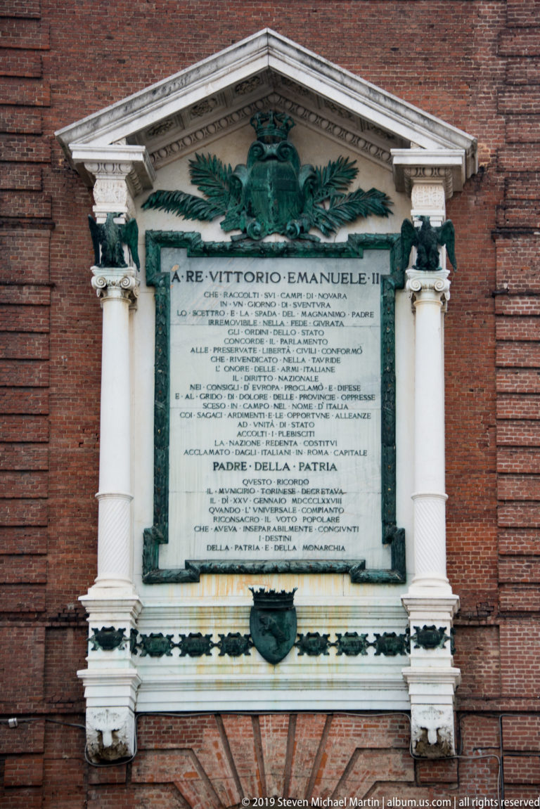 Ornate memorial plaque to Vittorio Emanuele II on a brick wall, framed by columns and a sculpted eagle.
