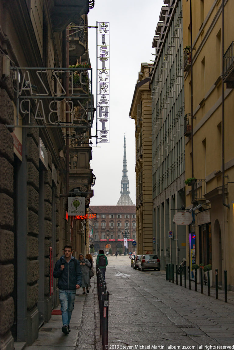 Straight Turin street lined with tall buildings and pedestrians, terminating in a distant view of the Mole Antonelliana spire.