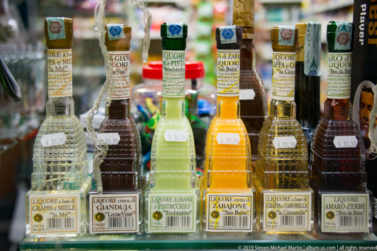 Rows of clear bottles containing differently colored Italian liqueurs, with flavor labels and prices visible.