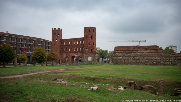 Distant Porta Palatina beyond a grassy archaeological field with low foundations and surviving ancient wall sections.