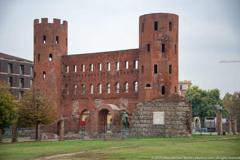Wide view of Porta Palatina with twin brick towers, arched openings, adjacent ancient walls, and grassy foreground.
