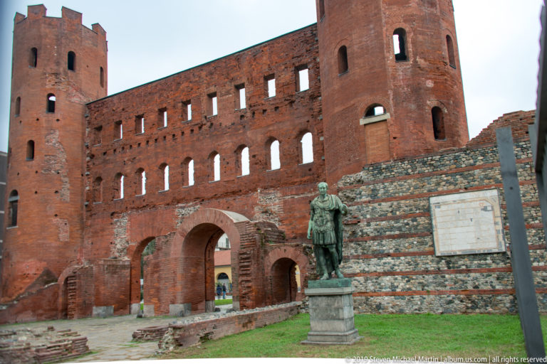 Oblique view of Porta Palatina’s brick tower and arches with a bronze standing statue in the foreground.