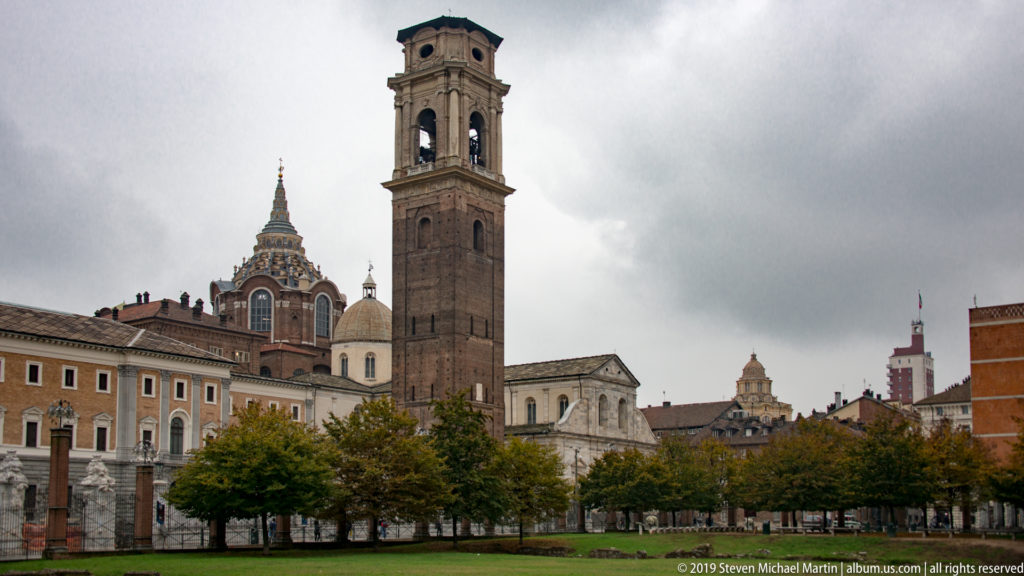 Tall historic bell tower above church roofs and pale buildings, with the Mole Antonelliana rising in the background.