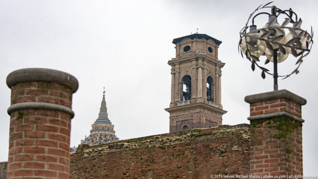 Brick archaeological walls below a square bell tower, with the Mole Antonelliana spire visible farther left.