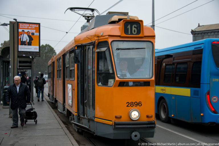 Orange route 16 tram stopped beside a blue bus, with passengers on the platform and overhead wires.