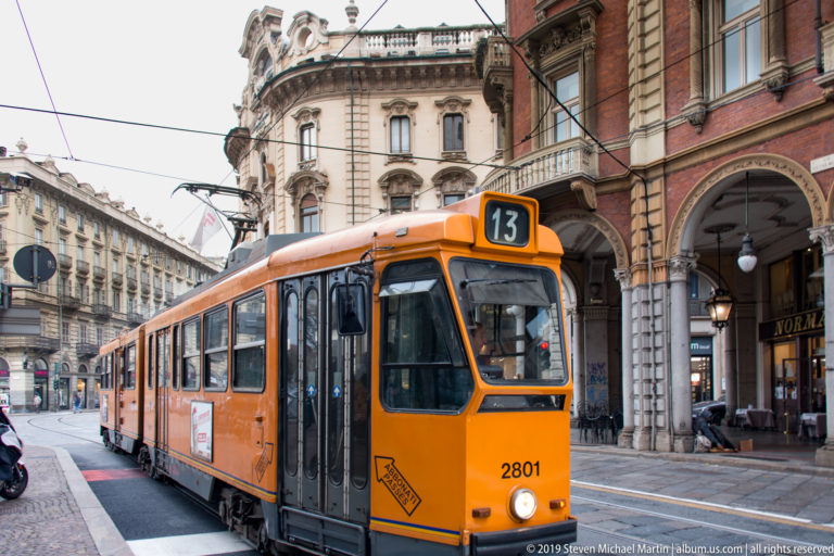 Orange route 13 tram number 2801 traveling on rails through a Turin street lined with arcaded historic buildings.