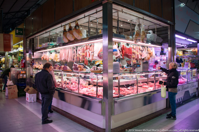 Customers at a brightly lit butcher counter displaying fresh meat, sausages, and hanging cured hams.