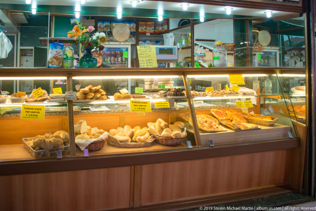 Glass market counter filled with baskets of bread, rolls, and trays of savory prepared foods with yellow labels.