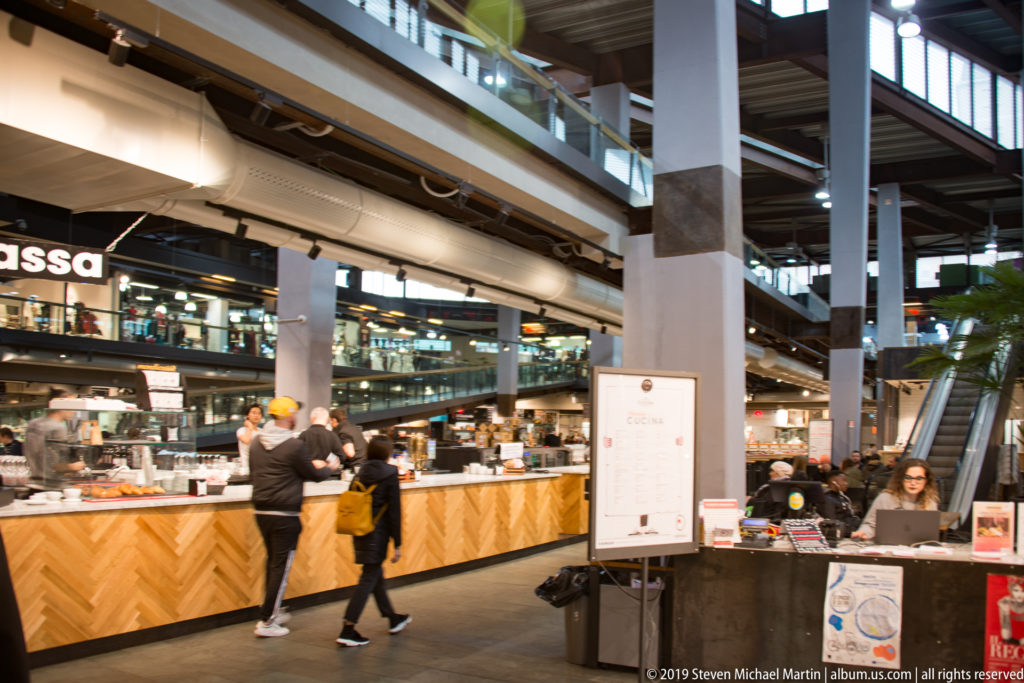 Shoppers at a wood-fronted food counter inside a multi-level market hall with vendors and seating beyond.