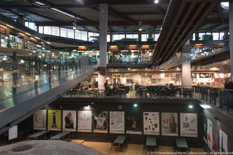 Multi-level indoor market with escalators, glass railings, food counters, tables, and exposed structural columns.