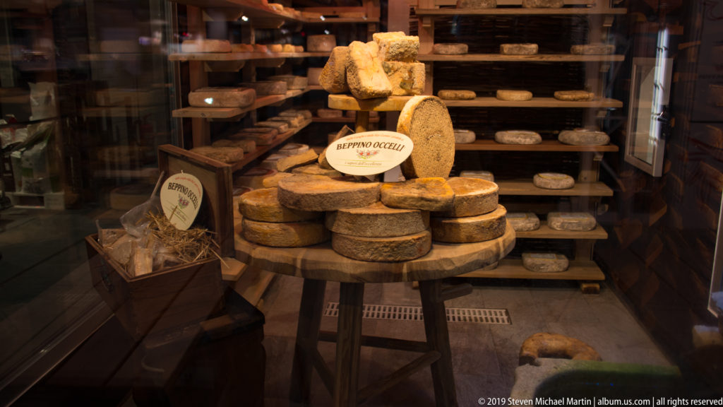 Rounds and wedges of aged cheese stacked on a market counter, with more cheese wheels on wooden shelves behind.
