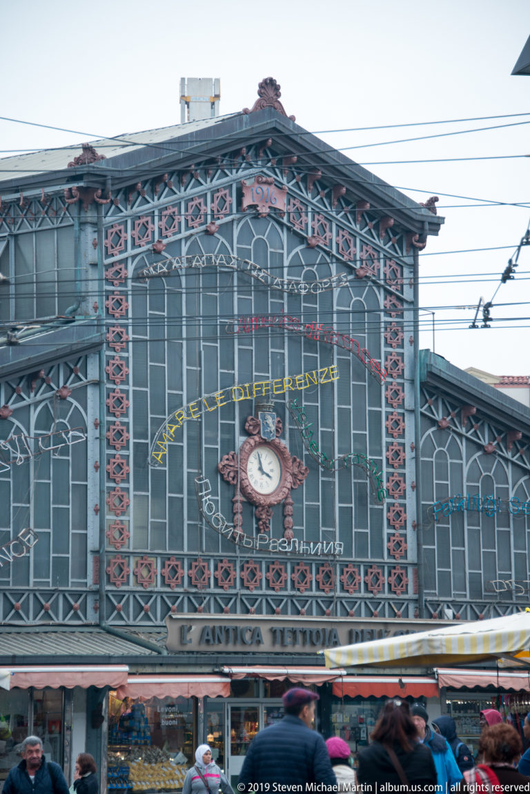 Iron-and-glass market hall with decorative red metalwork, central clock, market signage, stalls, and pedestrians.