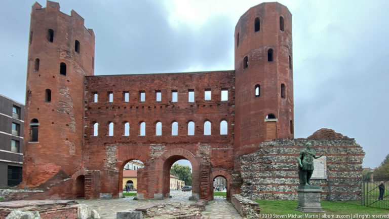 Red-brick Porta Palatina with twin towers, rows of arched openings, ground-level gateways, and adjacent ancient masonry.