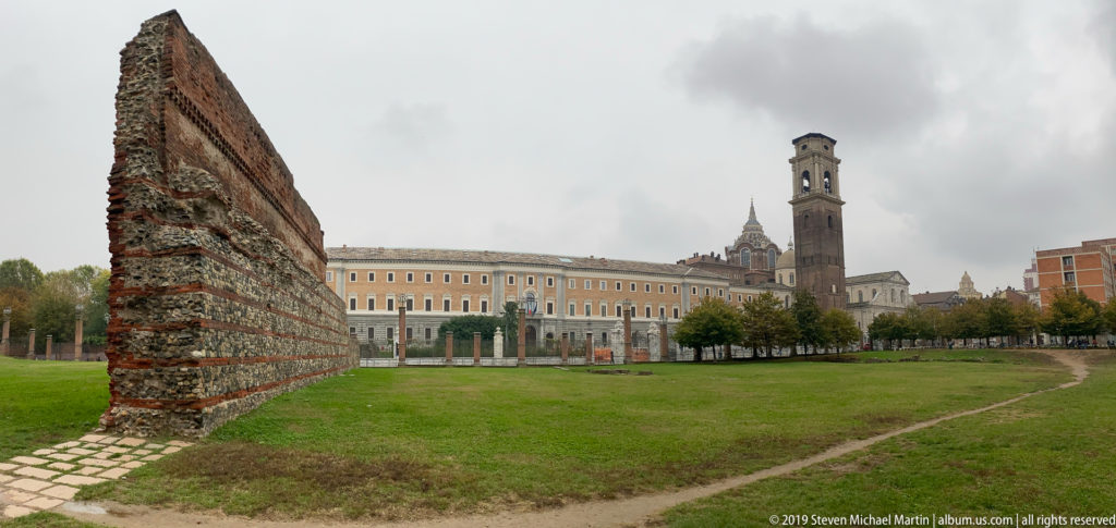 Ancient brick wall beside a grassy archaeological area, with historic buildings and a tall bell tower in the distance.
