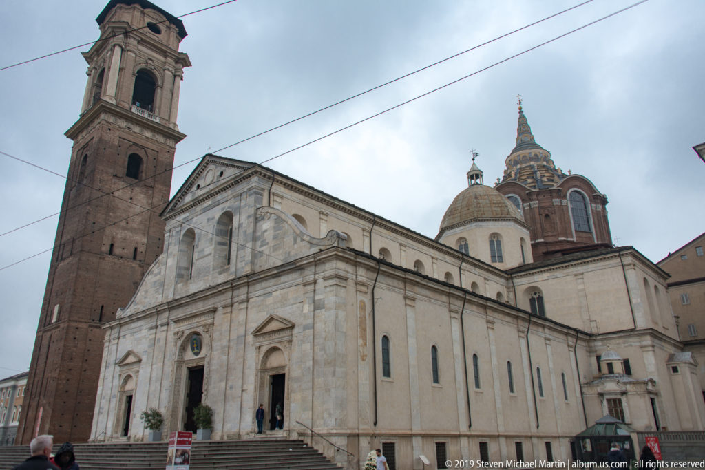 Oblique view of Turin Cathedral with pale stone façade, tall brick bell tower, and ornate Shroud Chapel dome rising behind.