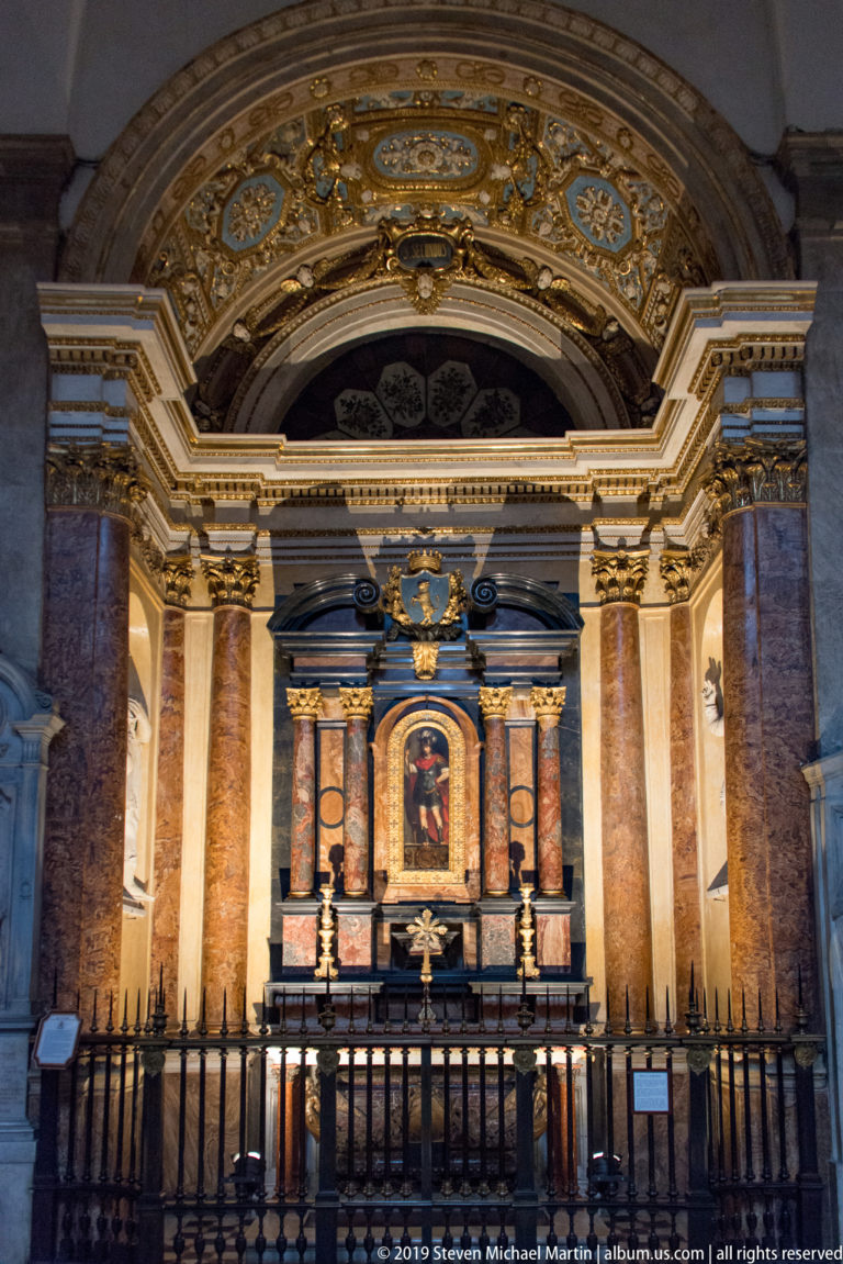 Symmetrical side chapel altar framed by paired colored-marble columns, gilded details, and a dark metal rail.