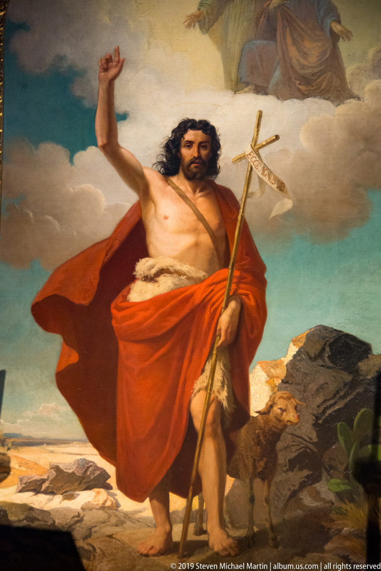 Saint John the Baptist stands in a rocky landscape wearing a red mantle and holding a cross-staff.