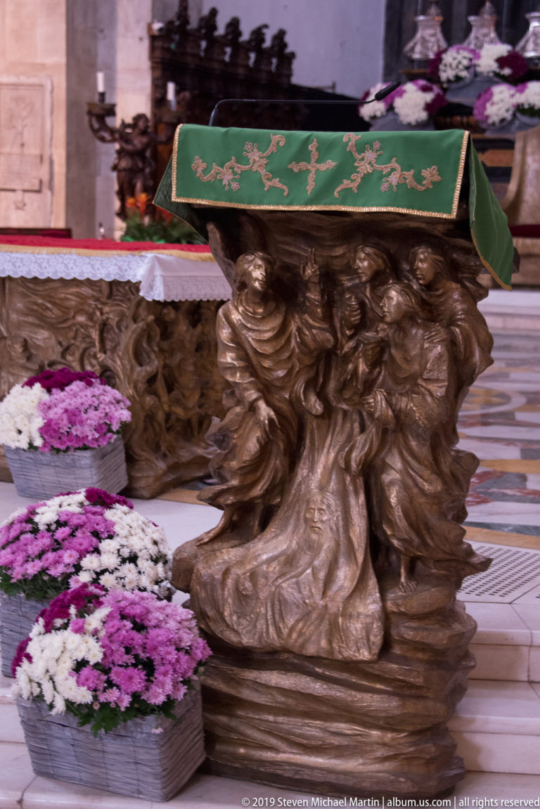 Side view of a figural altar support beneath a green cloth, with clustered sculpted bodies and devotional flowers nearby.