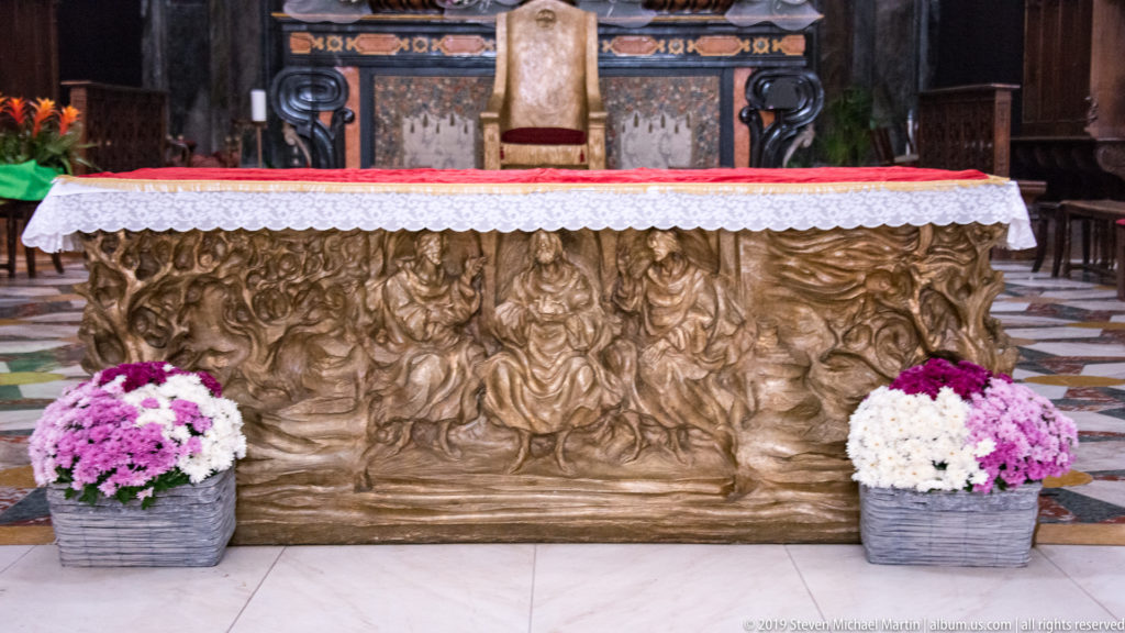 Bronze-toned altar frontal modeled with a dense group of figures, with flower pots placed symmetrically at both ends.