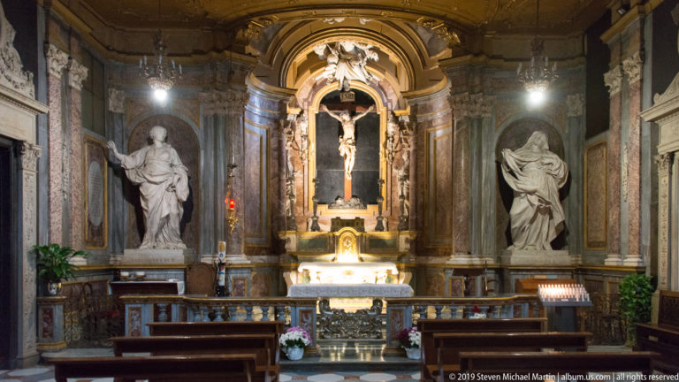 Crucifix centered above a chapel altar between two pale standing statues and dark marble architectural panels.