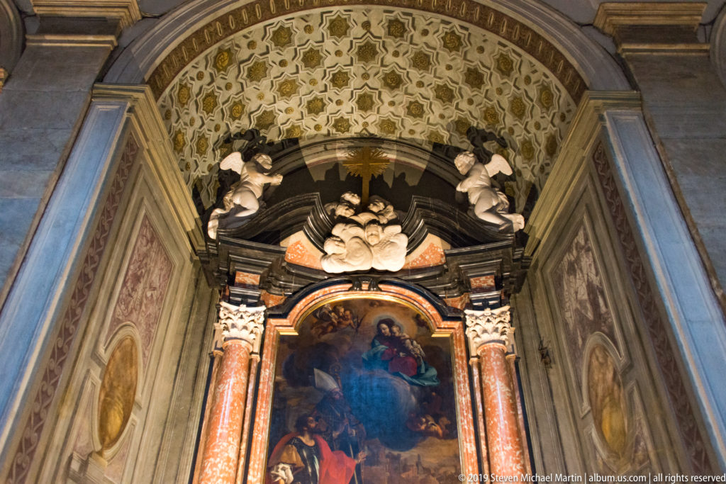 Upward view of a chapel altarpiece framed by colored marble columns, stucco figures, and a richly patterned vault.