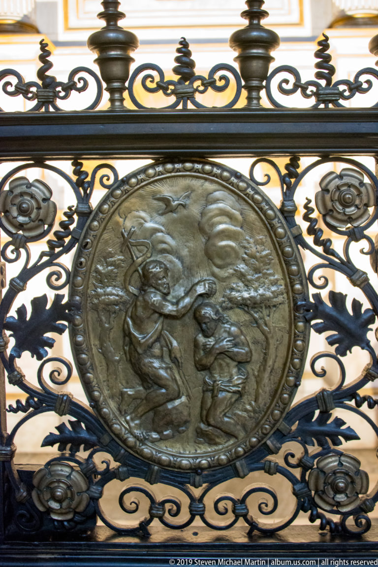 Circular figurative relief set within an elaborate wrought-metal grille of scrolling foliage and rosettes.