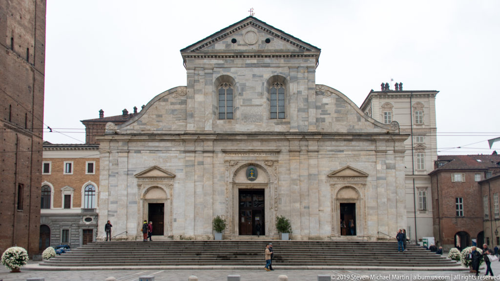 Symmetrical pale-stone façade of Turin Cathedral with three portals, broad steps, and a triangular pediment.