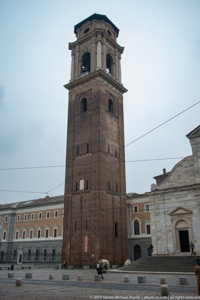 Tall square brick bell tower of Turin Cathedral rising from the piazza, with part of the cathedral façade at right.